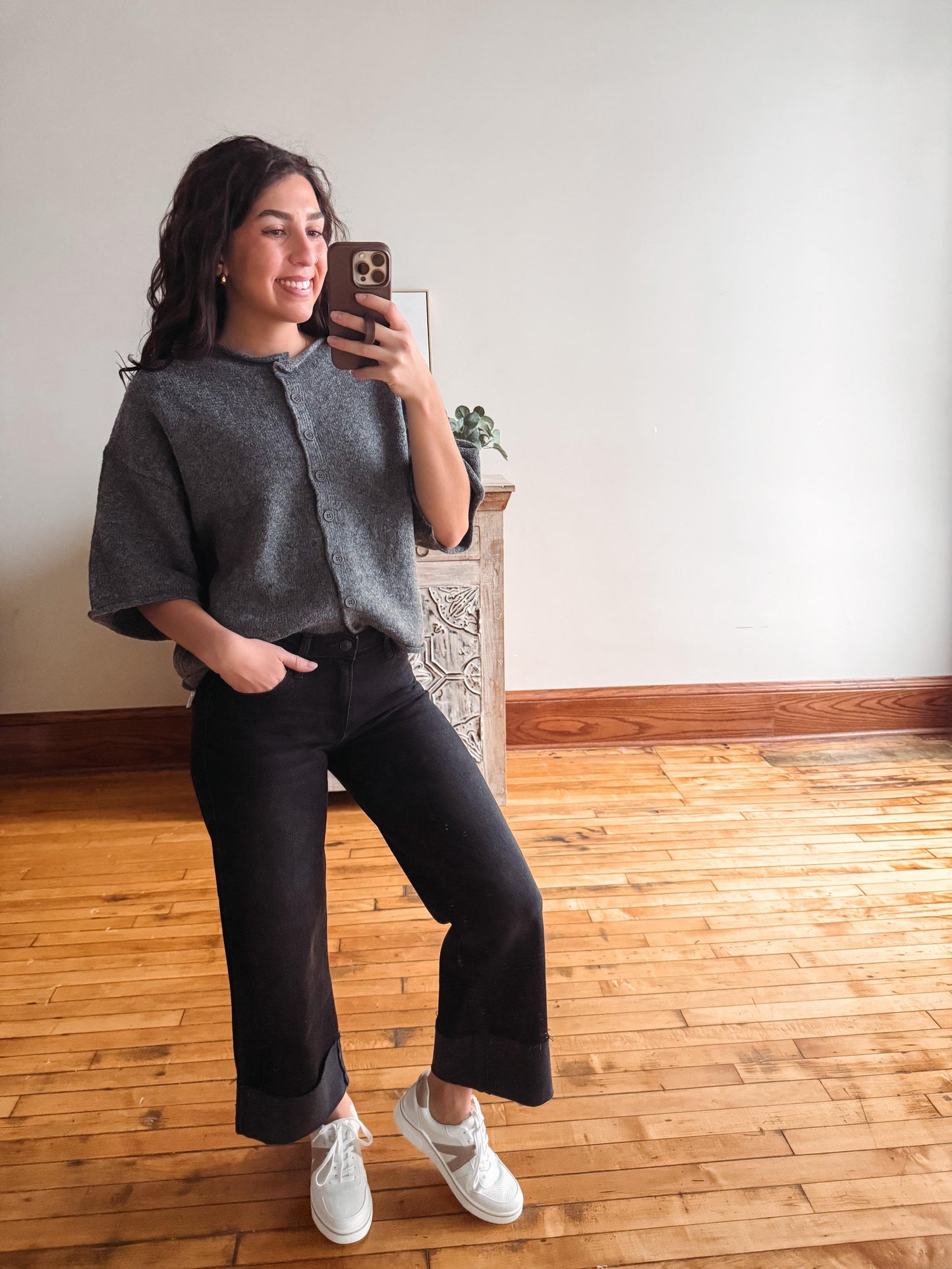 A woman takes a mirror selfie in a room with light wood floors and a white wall. She is wearing a gray textured short-sleeve top, black wide-leg cropped pants, and white sneakers.