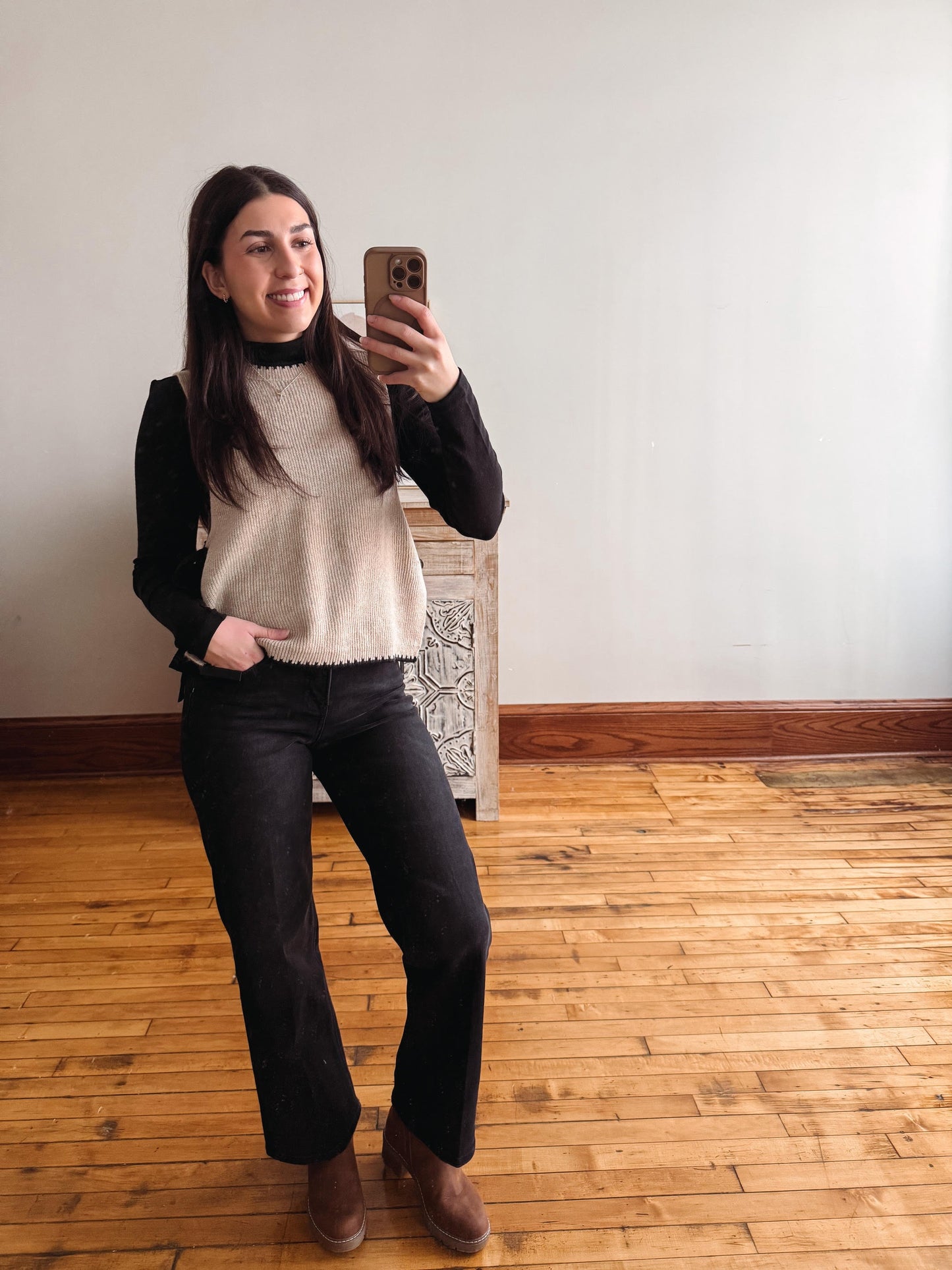 A woman takes a mirror selfie in a room with light-colored walls and wooden floors, wearing a cream knit sweater vest over a black long-sleeve top, black pants, and brown ankle boots.