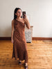 A woman takes a mirror selfie wearing a brown one-shoulder maxi dress with white polka dots, standing in a room with wooden floors and a white wall.