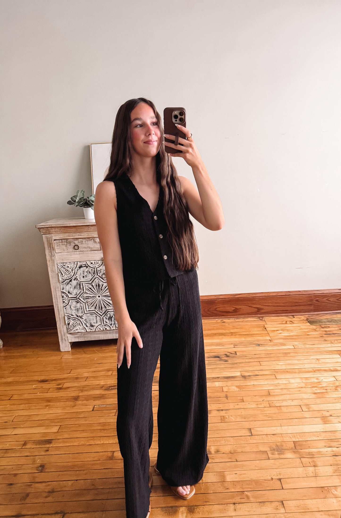 Person taking a mirror selfie wearing a black outfit in a room with wooden flooring and a decorative cabinet.