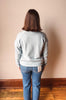 A person viewed from behind wearing a light heather gray knit sweater and blue jeans, standing against a plain beige wall with a wooden floor.