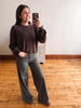 A woman takes a mirror selfie in a room with light wood floors and a white wall, wearing a dark brown ribbed knit sweater and dark olive green wide-leg pants.