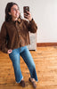 A woman takes a mirror selfie wearing a brown button-up shirt with lace detailing, blue jeans, and brown loafers in a room with wooden floors and a white wall.