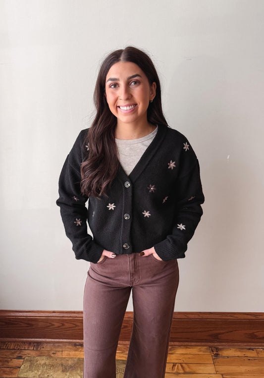 A woman stands indoors wearing a black knit cardigan with small white embroidered floral motifs, layered over a light gray top and paired with brown pants.