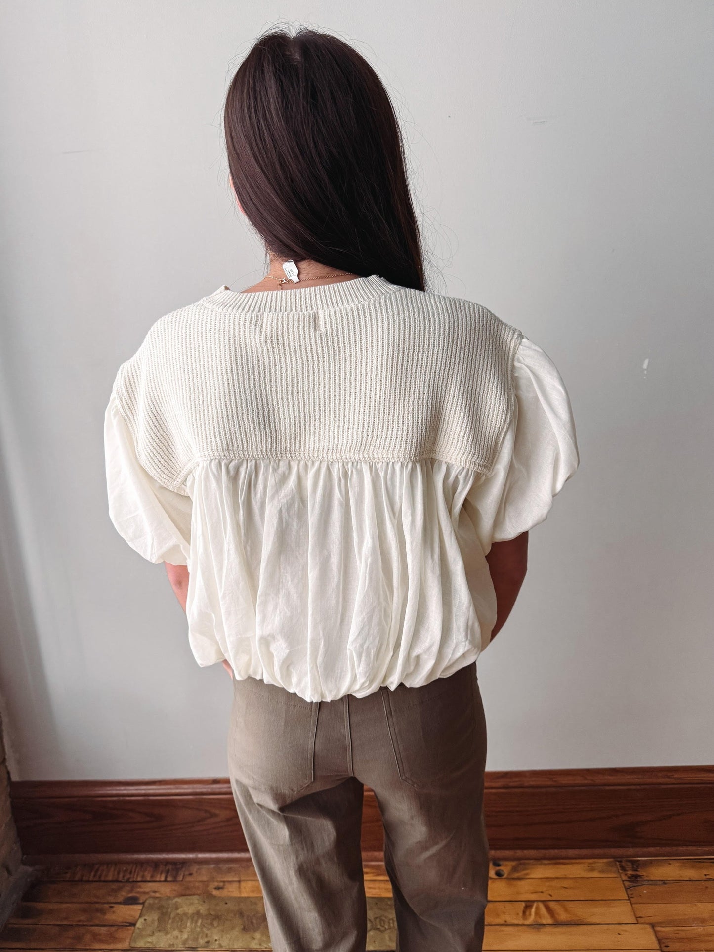 A woman viewed from behind wearing a cream-colored textured puff-sleeve blouse and taupe trousers, standing in front of a plain light gray wall with a wooden baseboard and floor.