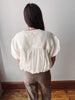 A woman viewed from behind wearing a cream-colored textured puff-sleeve blouse and taupe trousers, standing in front of a plain light gray wall with a wooden baseboard and floor.