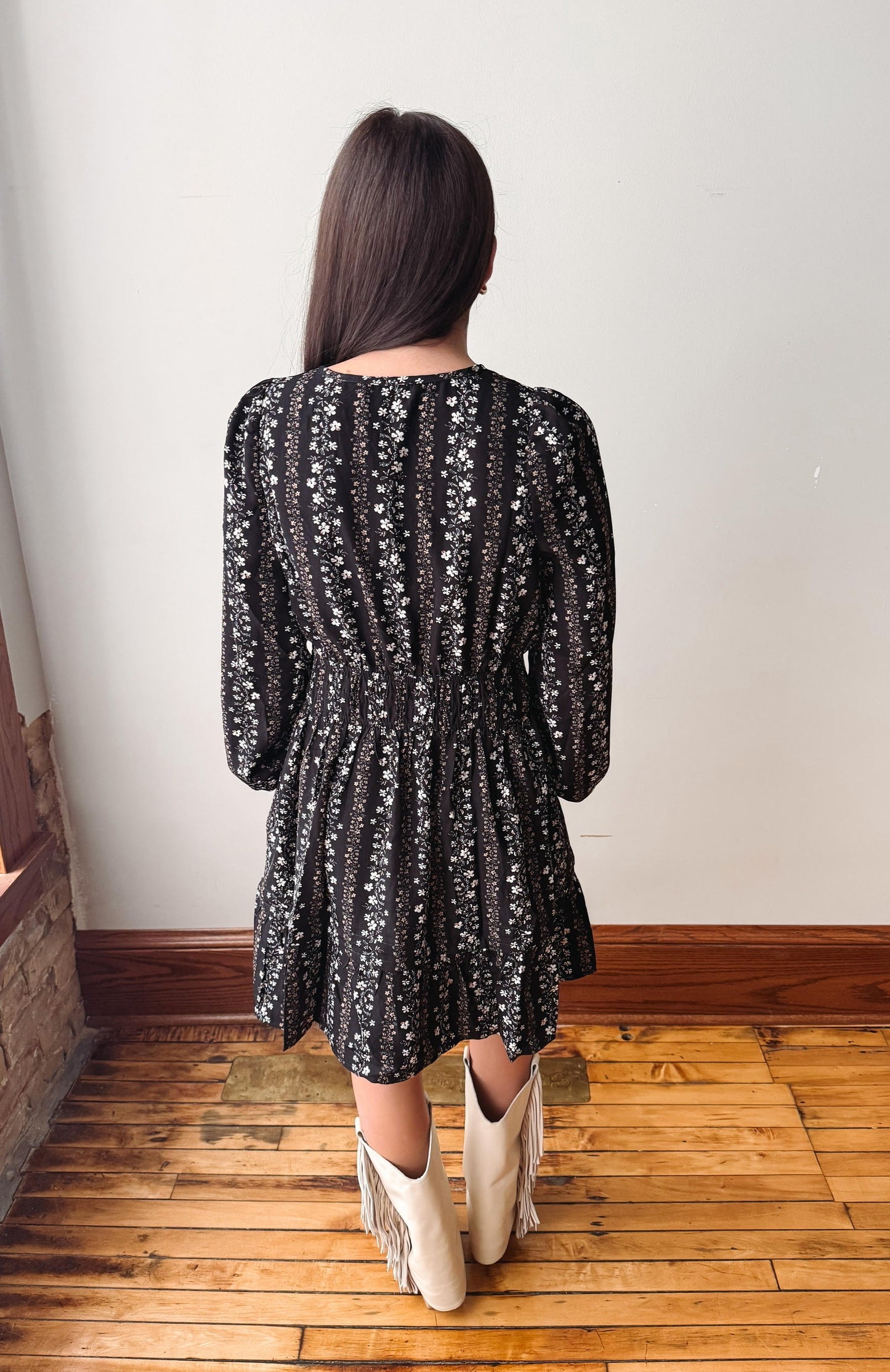 A woman viewed from behind wearing a black long-sleeve dress with a white floral pattern and cream fringe boots, standing on a wooden floor against a white wall.