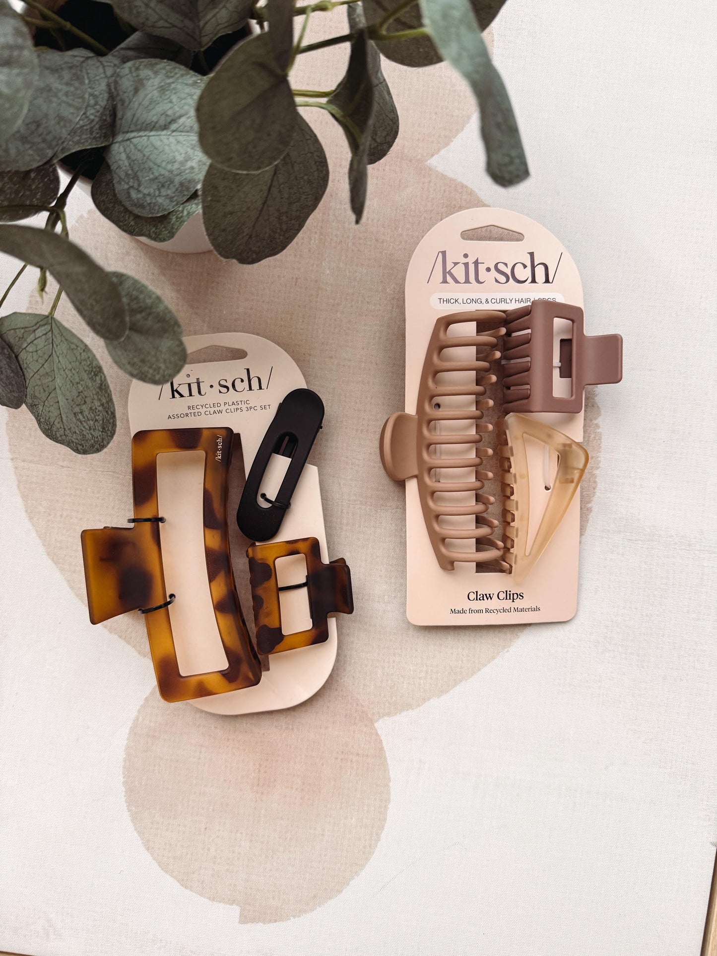 Two packaged hair claw clips on display cards, styled with eucalyptus leaves on a light fabric background. One set is tortoiseshell brown and black, and the other is taupe and beige.