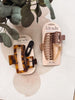 Two packaged hair claw clips on display cards, styled with eucalyptus leaves on a light fabric background. One set is tortoiseshell brown and black, and the other is taupe and beige.