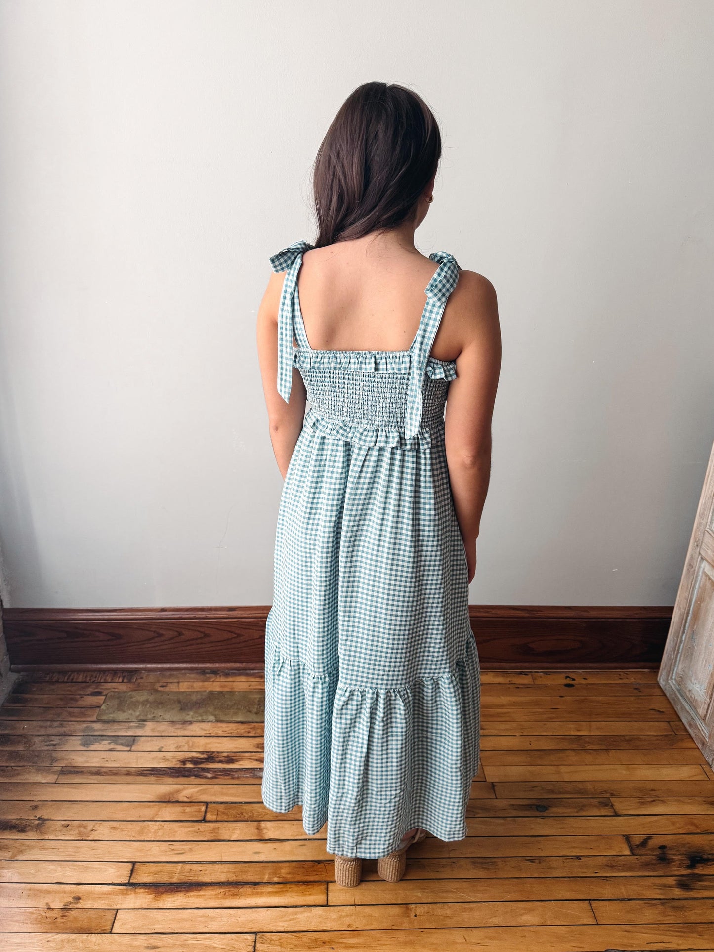 Woman wearing a green checkered dress standing on a wooden floor with a white wall background