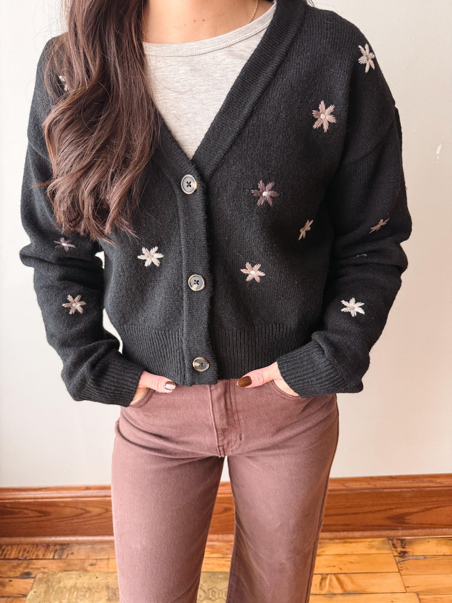 A person wearing a dark charcoal knit cardigan with light beige embroidered snowflake patterns, layered over a light gray top and paired with mauve-brown pants.