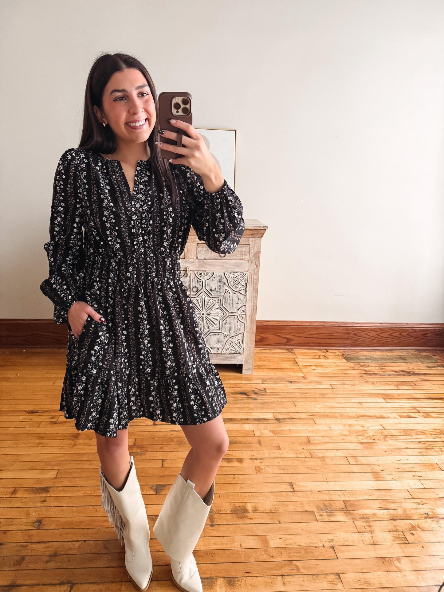 A woman takes a mirror selfie wearing a black long-sleeve mini dress with a white floral pattern and white cowboy boots, standing in a room with wooden floors and a white wall.