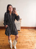 A woman takes a mirror selfie wearing a black long-sleeve mini dress with a white floral pattern and white cowboy boots, standing in a room with wooden floors and a white wall.