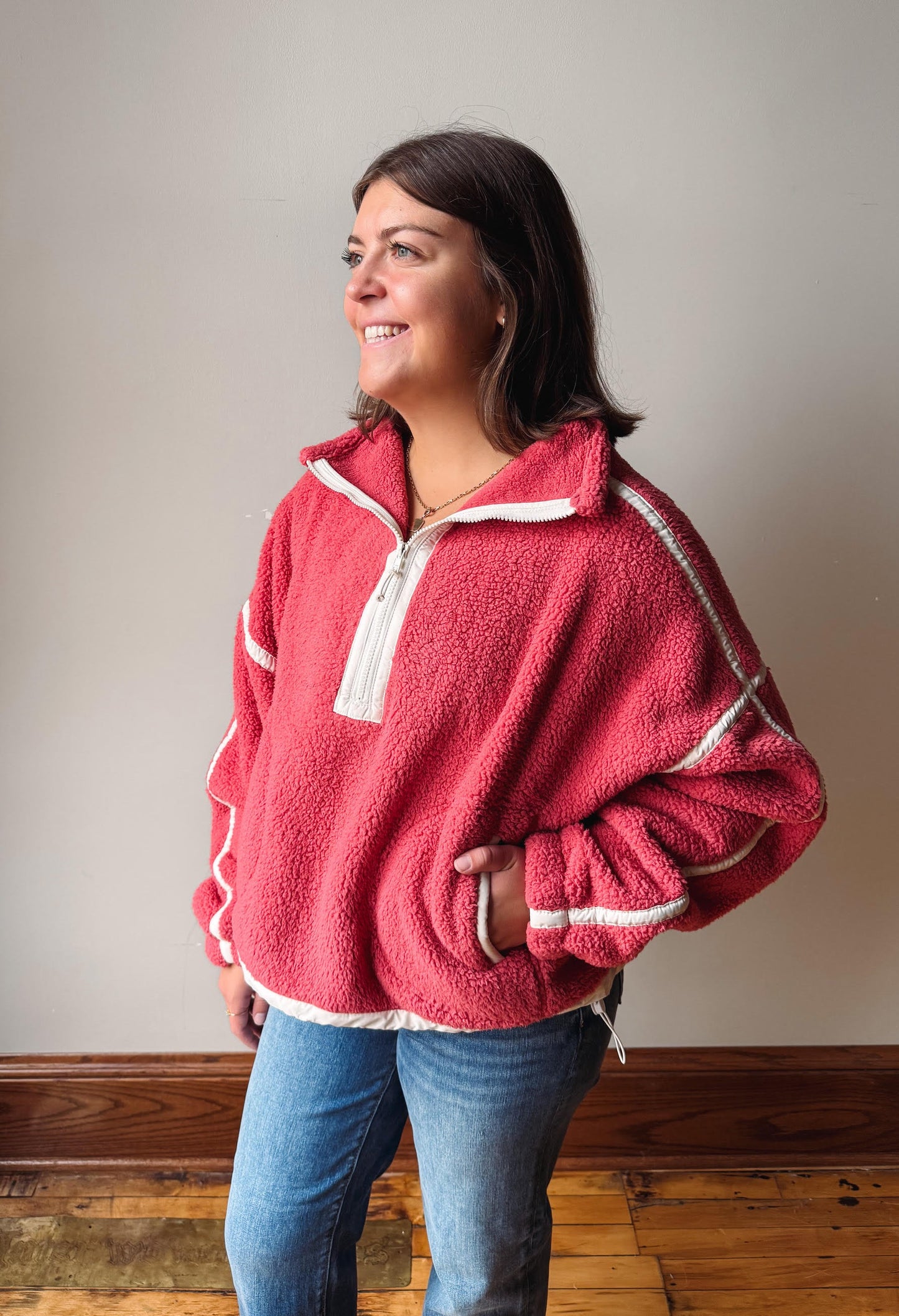 A woman wearing a dusty rose pink fleece pullover with white contrast trim and a white zipper, paired with blue jeans, standing against a neutral gray wall.