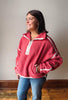 A woman wearing a dusty rose pink fleece pullover with white contrast trim and a white zipper, paired with blue jeans, standing against a neutral gray wall.