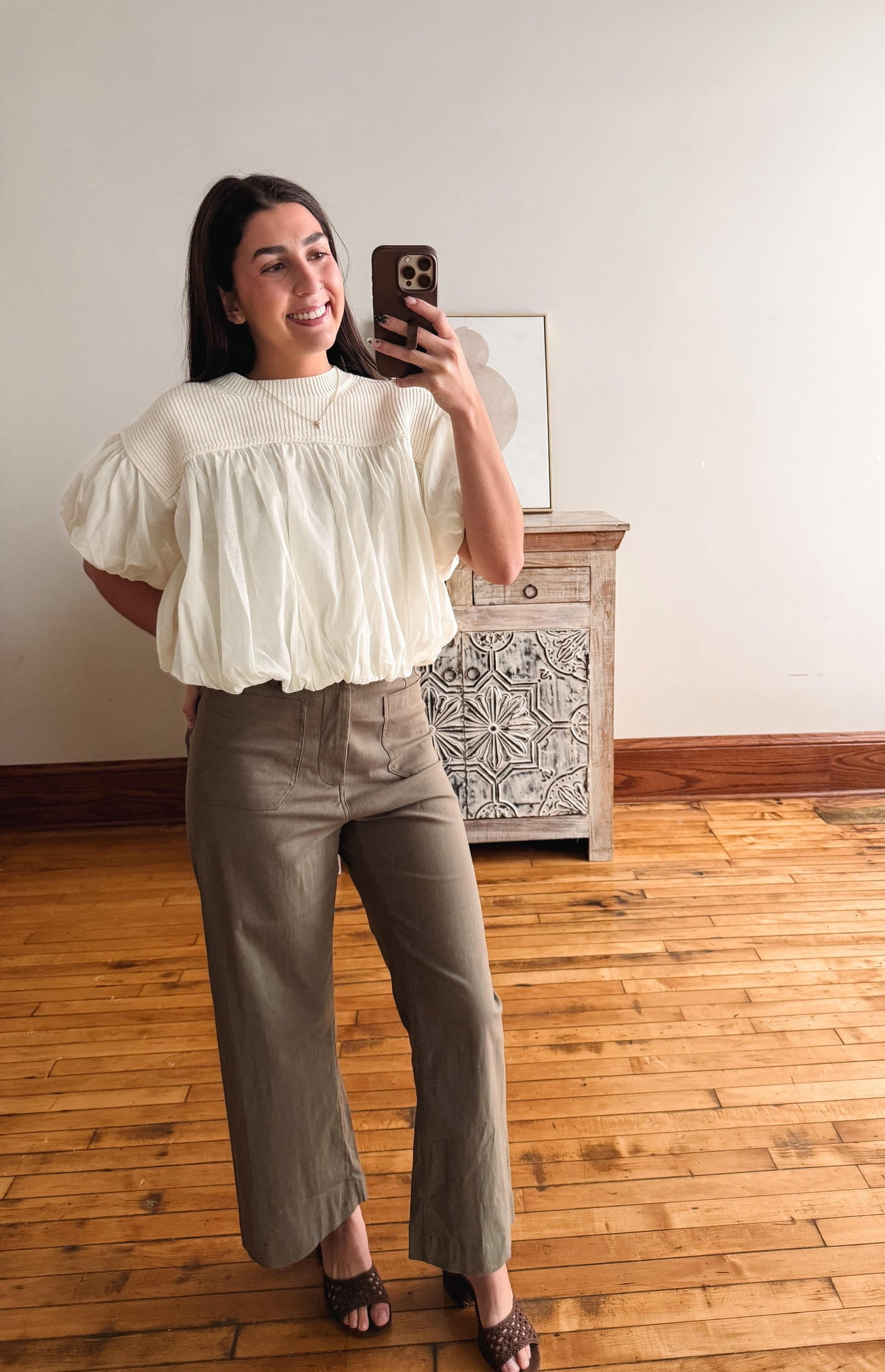 A woman takes a mirror selfie in a room with light wood floors and a white wall, wearing a cream puff-sleeve blouse and olive green wide-leg pants.