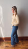 Woman wearing a yellow sweater and blue jeans standing against a white wall.