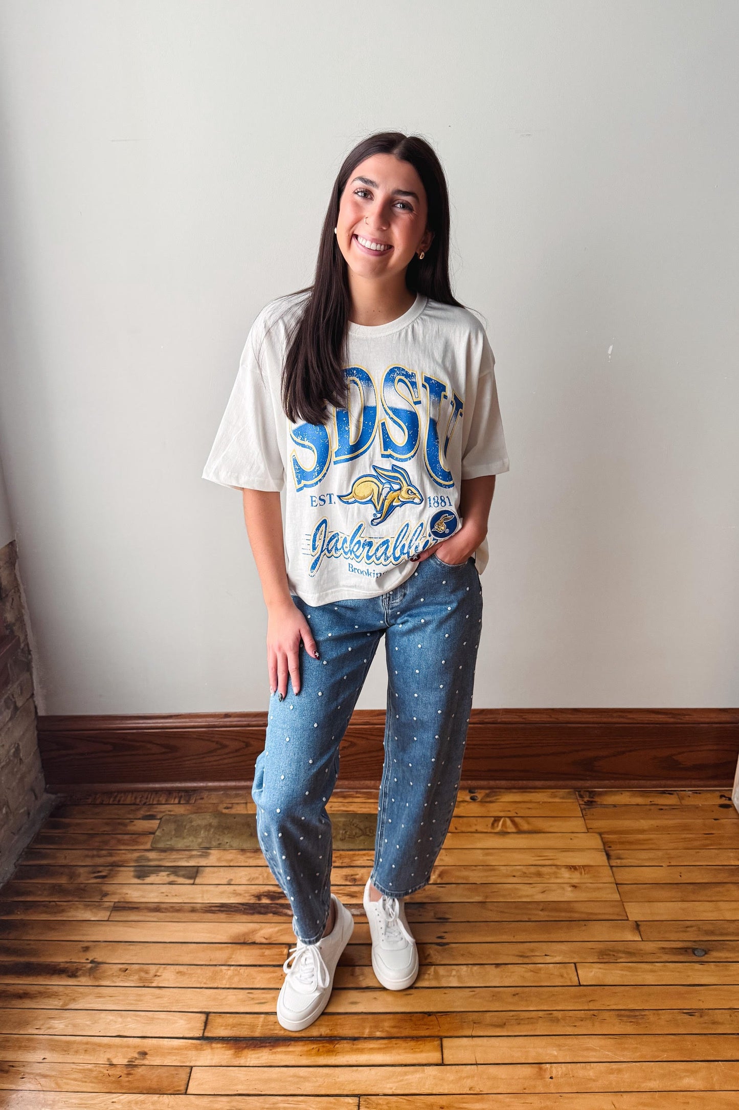 A woman modeling a white graphic t-shirt with blue lettering that reads 'DSU' and 'Guelaguetza', paired with blue jeans featuring a white speckled pattern and white sneakers, standing on a wooden floor against a plain wall.
