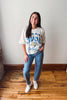 A woman modeling a white graphic t-shirt with blue lettering that reads 'DSU' and 'Guelaguetza', paired with blue jeans featuring a white speckled pattern and white sneakers, standing on a wooden floor against a plain wall.