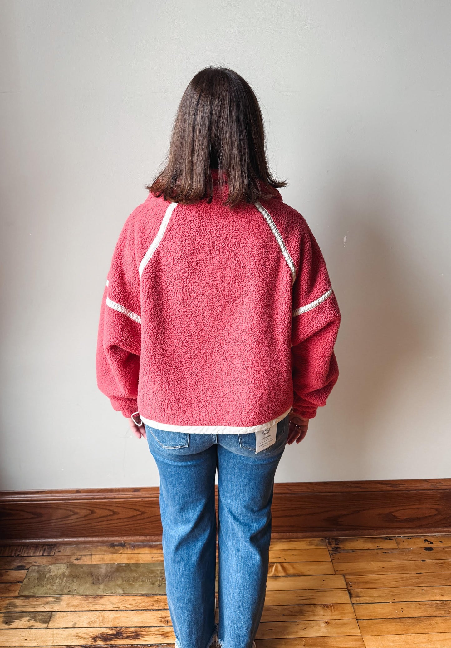 A person viewed from behind wearing a pink textured fleece zip-up jacket with white contrast stitching and blue jeans, standing on a wooden floor against a plain light gray wall.