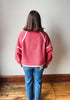 A person viewed from behind wearing a pink textured fleece zip-up jacket with white contrast stitching and blue jeans, standing on a wooden floor against a plain light gray wall.