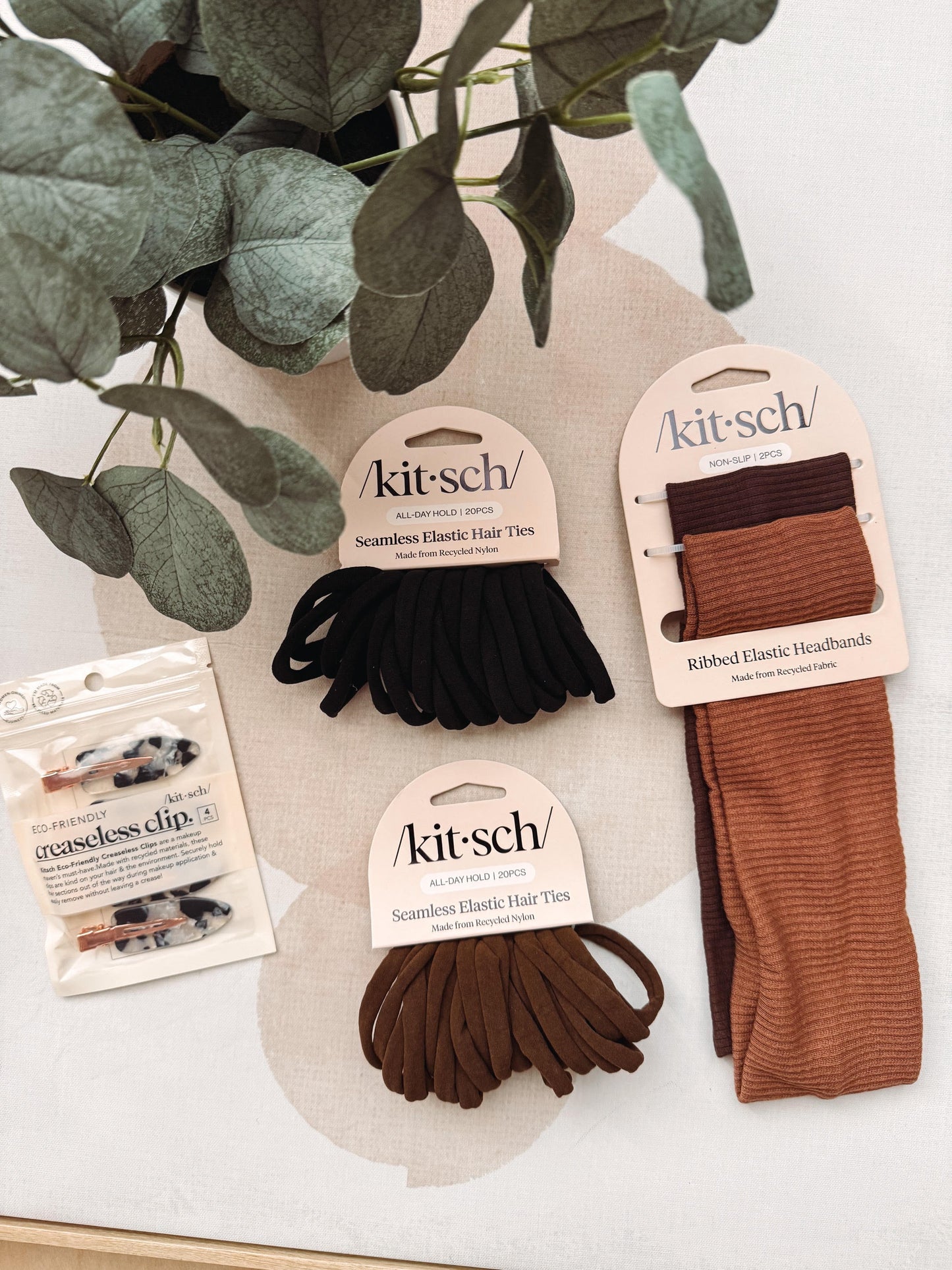 A Kit Sch collection hair accessory set displayed on a light beige surface with a green plant in the background. The set includes three packages of elastic hair ties in black, brown, and rust, along with a pack of marble-patterned hair clips.