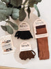 A Kit Sch collection hair accessory set displayed on a light beige surface with a green plant in the background. The set includes three packages of elastic hair ties in black, brown, and rust, along with a pack of marble-patterned hair clips.
