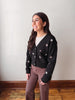 A woman wearing a black knit cardigan with white embroidered star patterns, styled with brown pants and a white top, standing against a plain off-white wall.