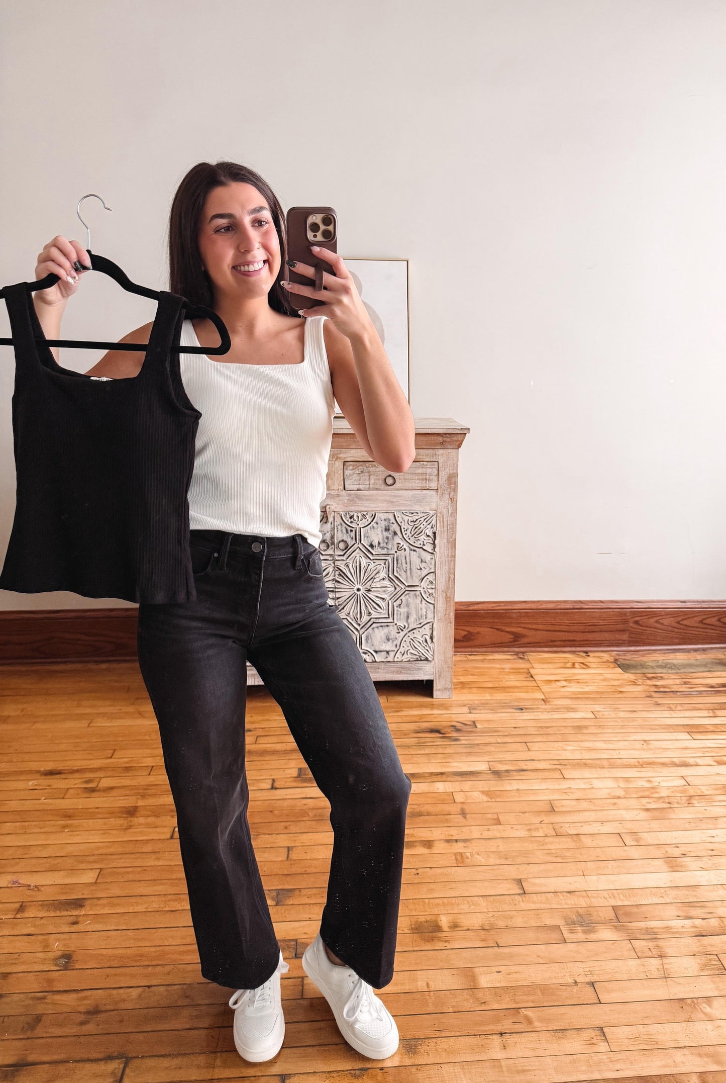 A woman takes a mirror selfie in a room with hardwood floors and a white wall, holding up a black sleeveless top on a hanger next to her white tank top and black jeans.