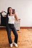 A woman takes a mirror selfie in a room with hardwood floors and a white wall, holding up a black sleeveless top on a hanger next to her white tank top and black jeans.
