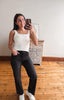 A woman takes a mirror selfie wearing a white ribbed tank top, black wide-leg jeans, and white sneakers in a room with light-colored walls and wooden floors.
