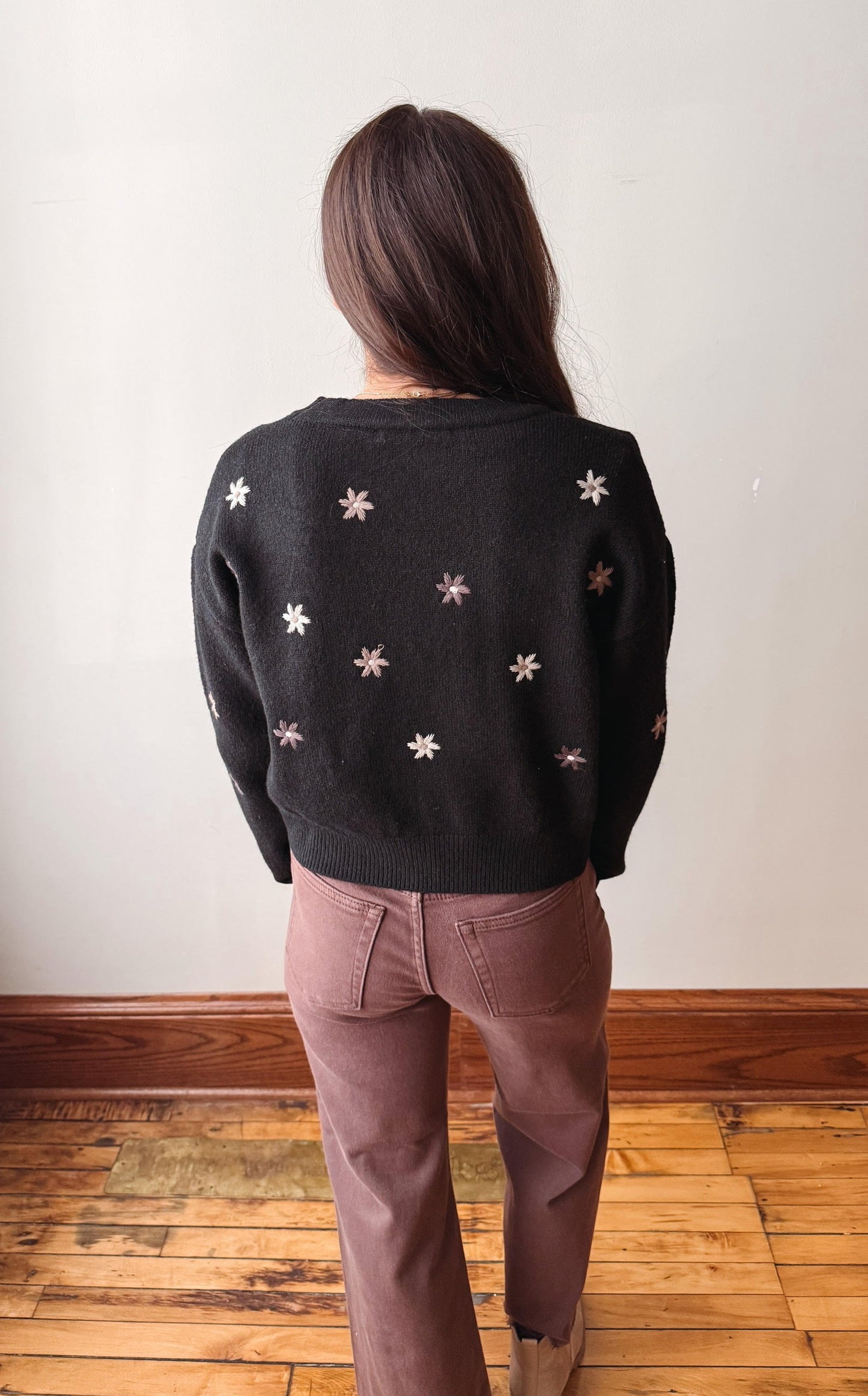 A woman viewed from behind wearing a black knit sweater with scattered white embroidered star motifs and mauve-brown pants, standing on a wooden floor against a plain off-white wall.