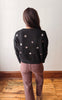A woman viewed from behind wearing a black knit sweater with scattered white embroidered star motifs and mauve-brown pants, standing on a wooden floor against a plain off-white wall.