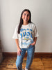 A woman stands indoors wearing a cream-colored vintage-style graphic t-shirt with blue and yellow 'SOUTH DAKOTA JAYALITHS' lettering, paired with blue jeans featuring a white star pattern.
