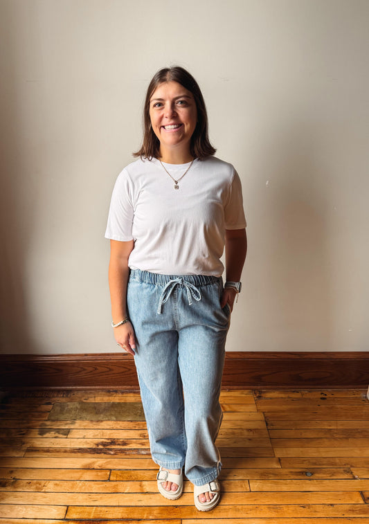A woman stands in a room with a beige wall and wooden floor, wearing a white short-sleeve t-shirt, light-wash blue drawstring sweatpants, and white slide sandals.