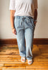 A person wearing light-wash blue drawstring jeans, a white t-shirt, and white slide sandals, standing on a wooden floor against a plain wall.