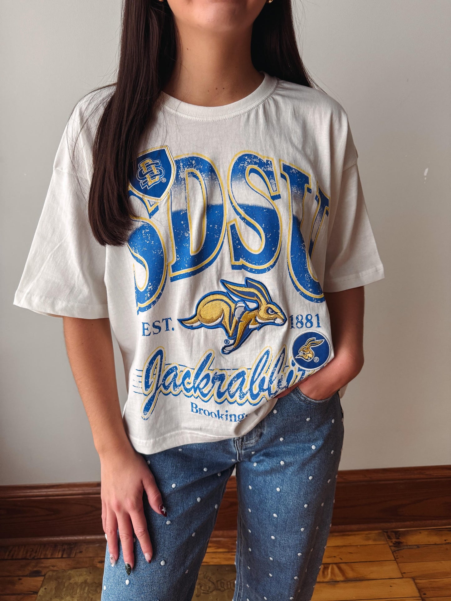 A person wearing an oversized vintage-style white graphic T-shirt with large blue and yellow 'SDSU' lettering, a kangaroo graphic, and 'JACKABRIDGE BROOKINGS' text, paired with blue jeans with small white dots, standing indoors against a plain wall.