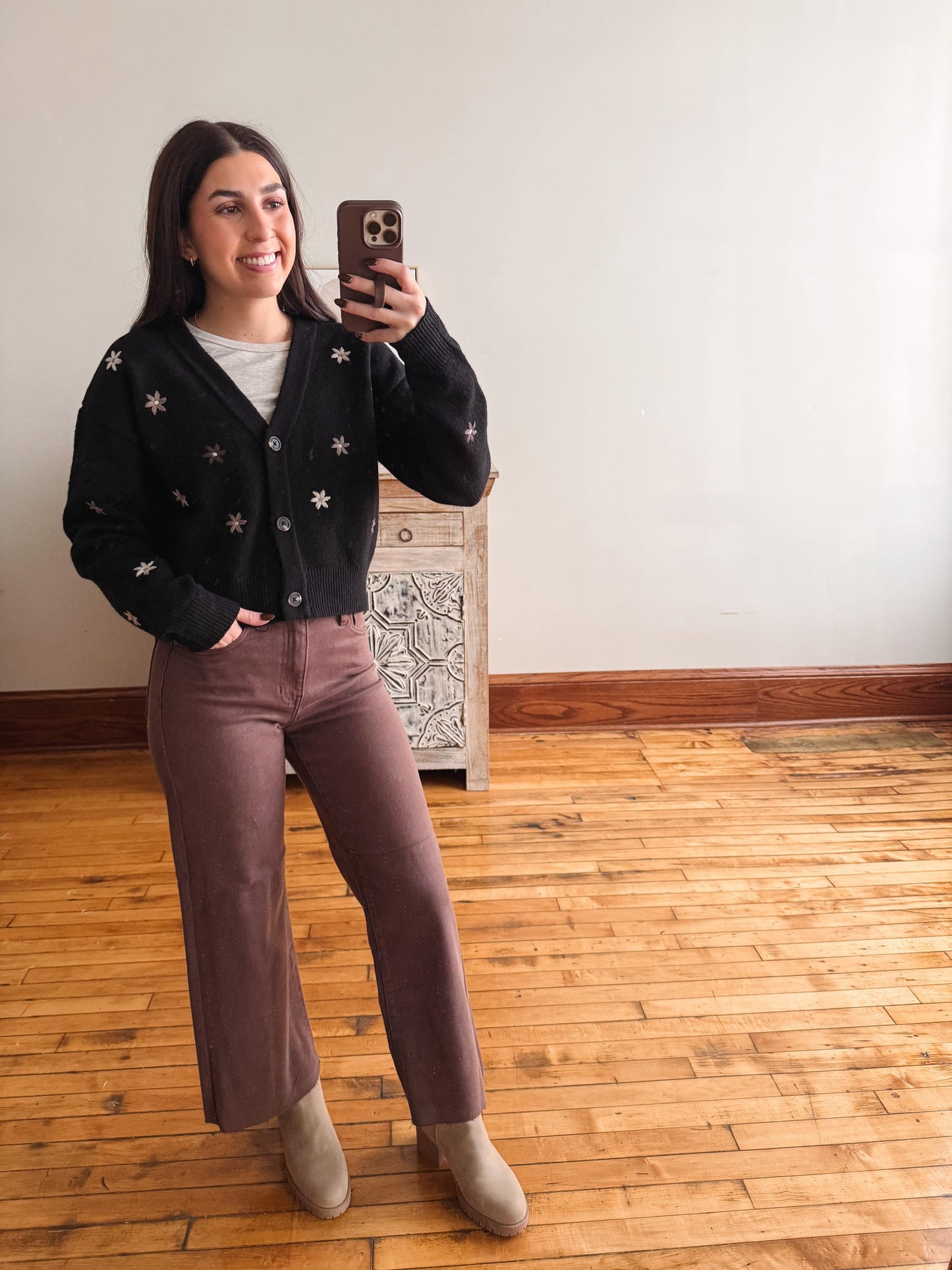 A woman takes a mirror selfie in a room with hardwood floors and a white wall, wearing a black button-up cardigan with white embroidered stars, mauve wide-leg pants, and beige ankle boots.