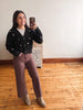 A woman takes a mirror selfie in a room with hardwood floors and a white wall, wearing a black button-up cardigan with white embroidered stars, mauve wide-leg pants, and beige ankle boots.