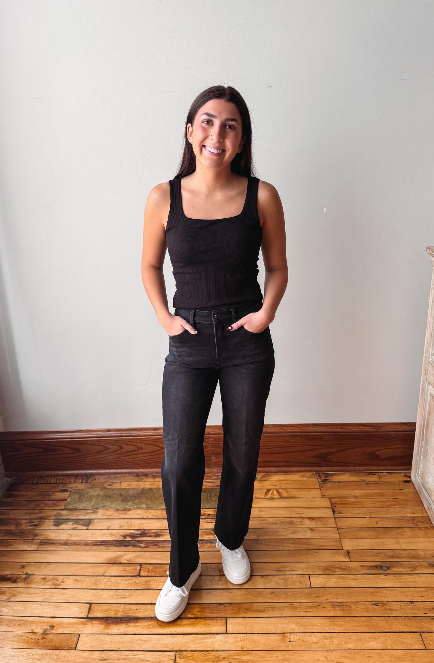 A woman stands in a room with a white wall and wooden floor, wearing a black tank top, black jeans, and white sneakers.