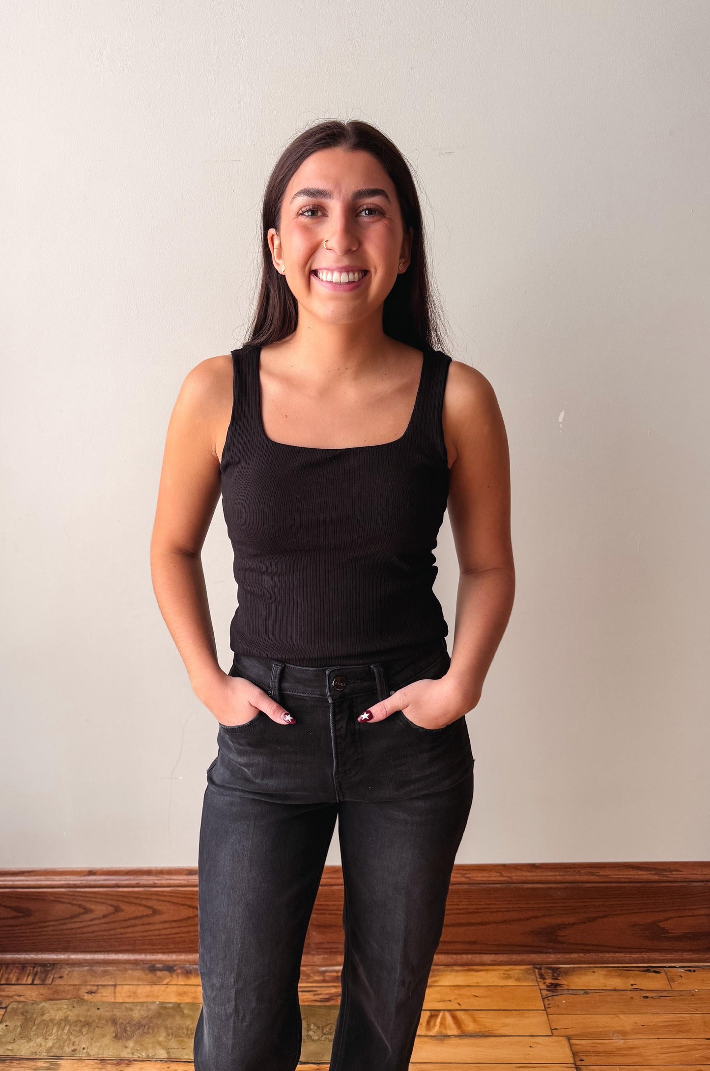 A smiling young woman with long dark hair stands indoors wearing a black sleeveless tank top and dark jeans, with her hands in her pockets, against a plain light wall and wood floor.