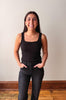 A smiling young woman with long dark hair stands indoors wearing a black sleeveless tank top and dark jeans, with her hands in her pockets, against a plain light wall and wood floor.