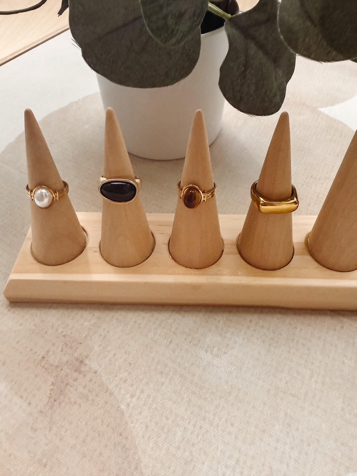 Five gold-tone ring stands made of light wood are arranged on a matching rectangular base, each holding a ring with different stone colors, set on a light fabric surface with a potted plant in the background.