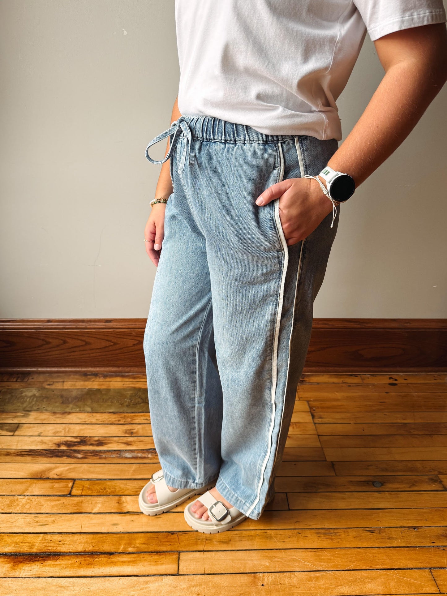 A person wearing light-wash wide-leg denim jeans with a drawstring waist, styled with a white t-shirt and white slide sandals, standing on a wooden floor against a neutral wall.