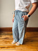 A person wearing light-wash wide-leg denim jeans with a drawstring waist, styled with a white t-shirt and white slide sandals, standing on a wooden floor against a neutral wall.