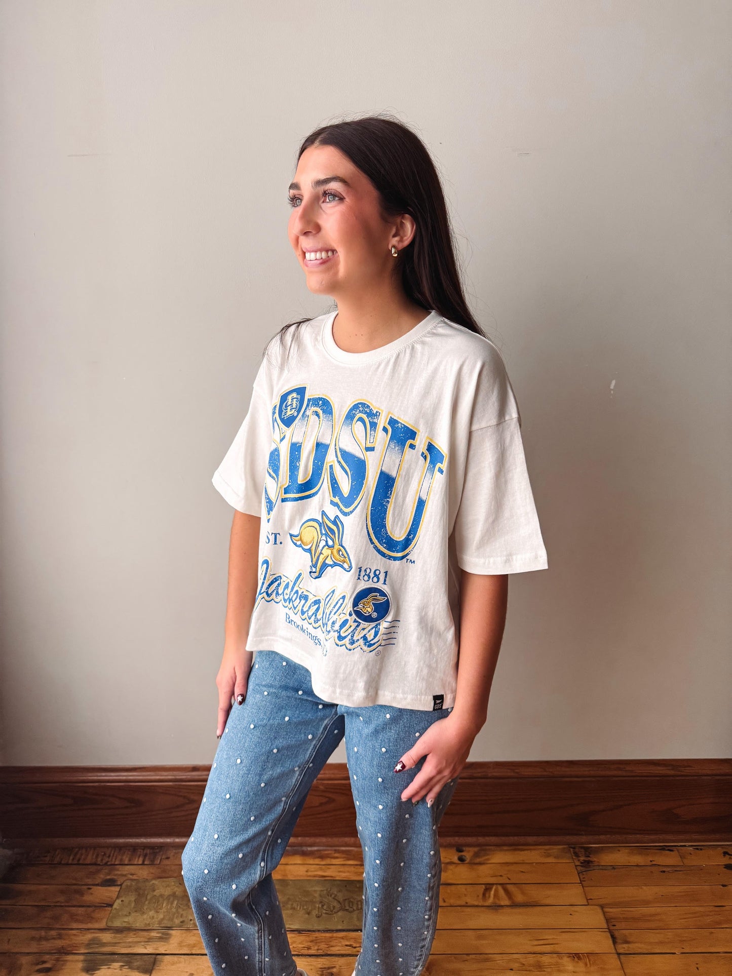 A woman modeling a vintage off-white short-sleeve t-shirt with a blue and yellow 'LSU' graphic, paired with blue jeans, standing in front of a plain beige wall and wooden floor.