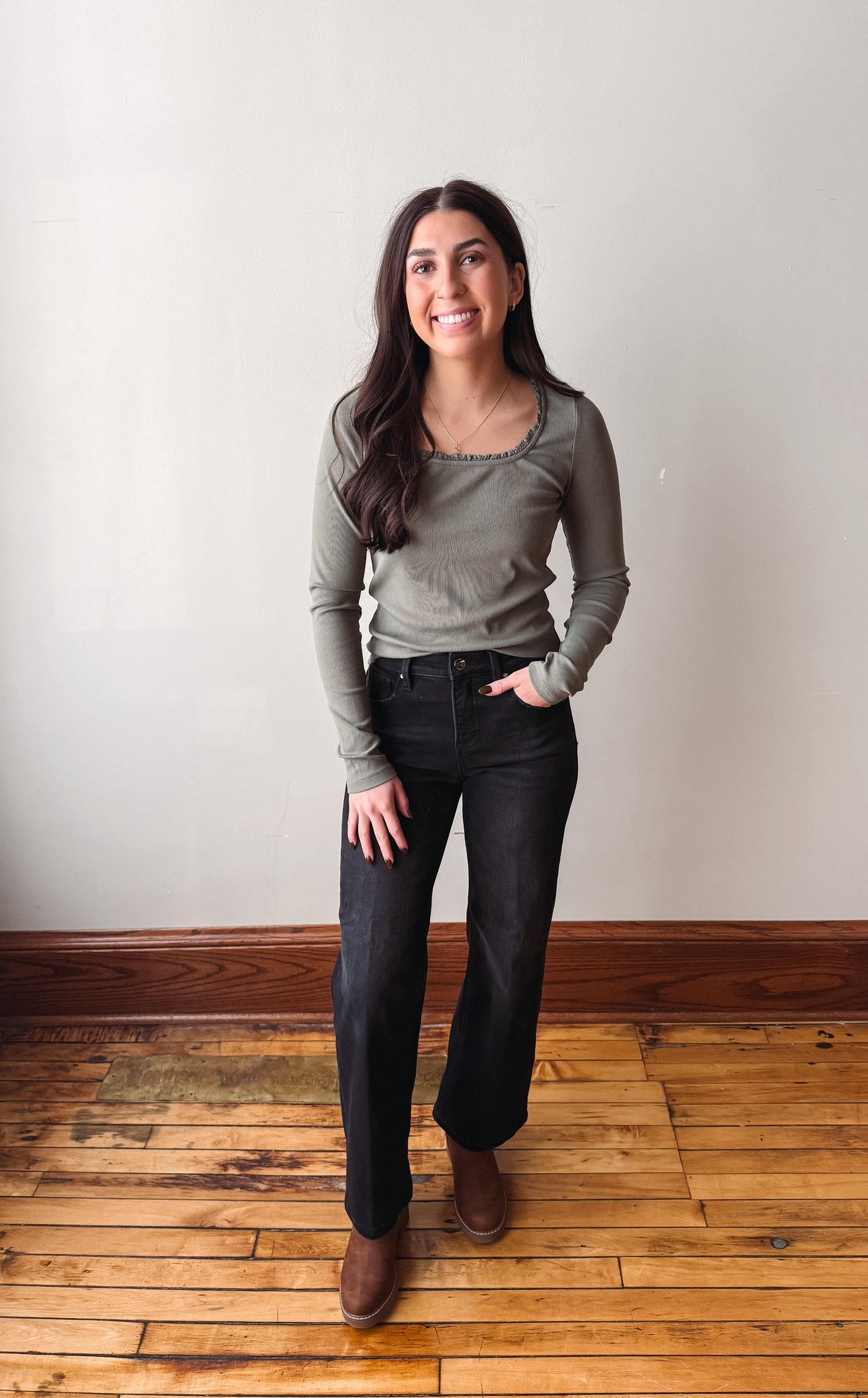 A woman stands in a room with a white wall and wooden floor, wearing a ribbed long-sleeve top in a muted sage green, black wide-leg jeans, and brown boots.