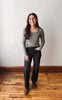 A woman stands in a room with a white wall and wooden floor, wearing a ribbed long-sleeve top in a muted sage green, black wide-leg jeans, and brown boots.