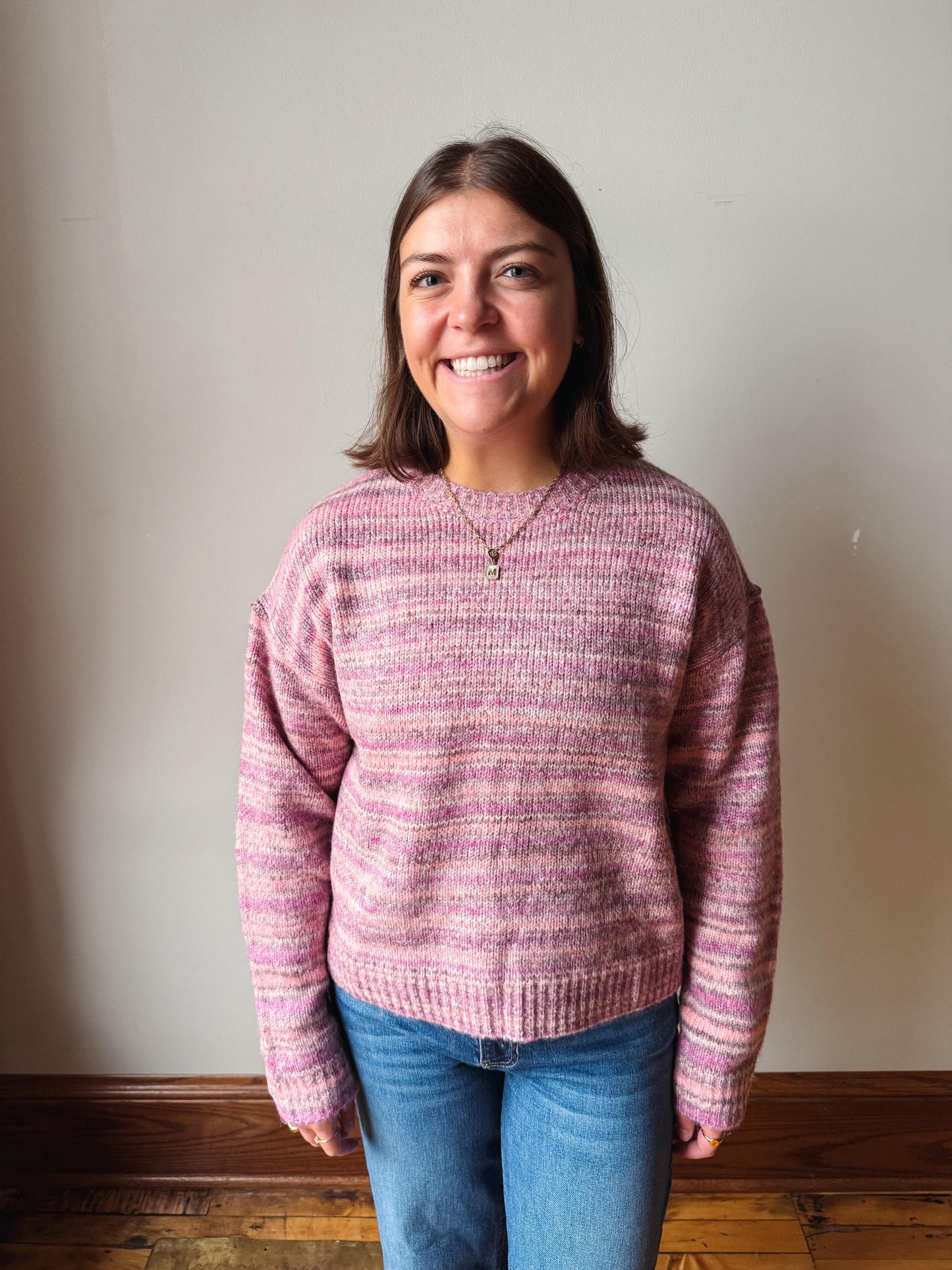 A smiling woman with shoulder-length brown hair stands indoors wearing a pink and mauve striped knit sweater and blue jeans against a plain off-white wall.