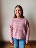 A smiling woman with shoulder-length brown hair stands indoors wearing a pink and mauve striped knit sweater and blue jeans against a plain off-white wall.